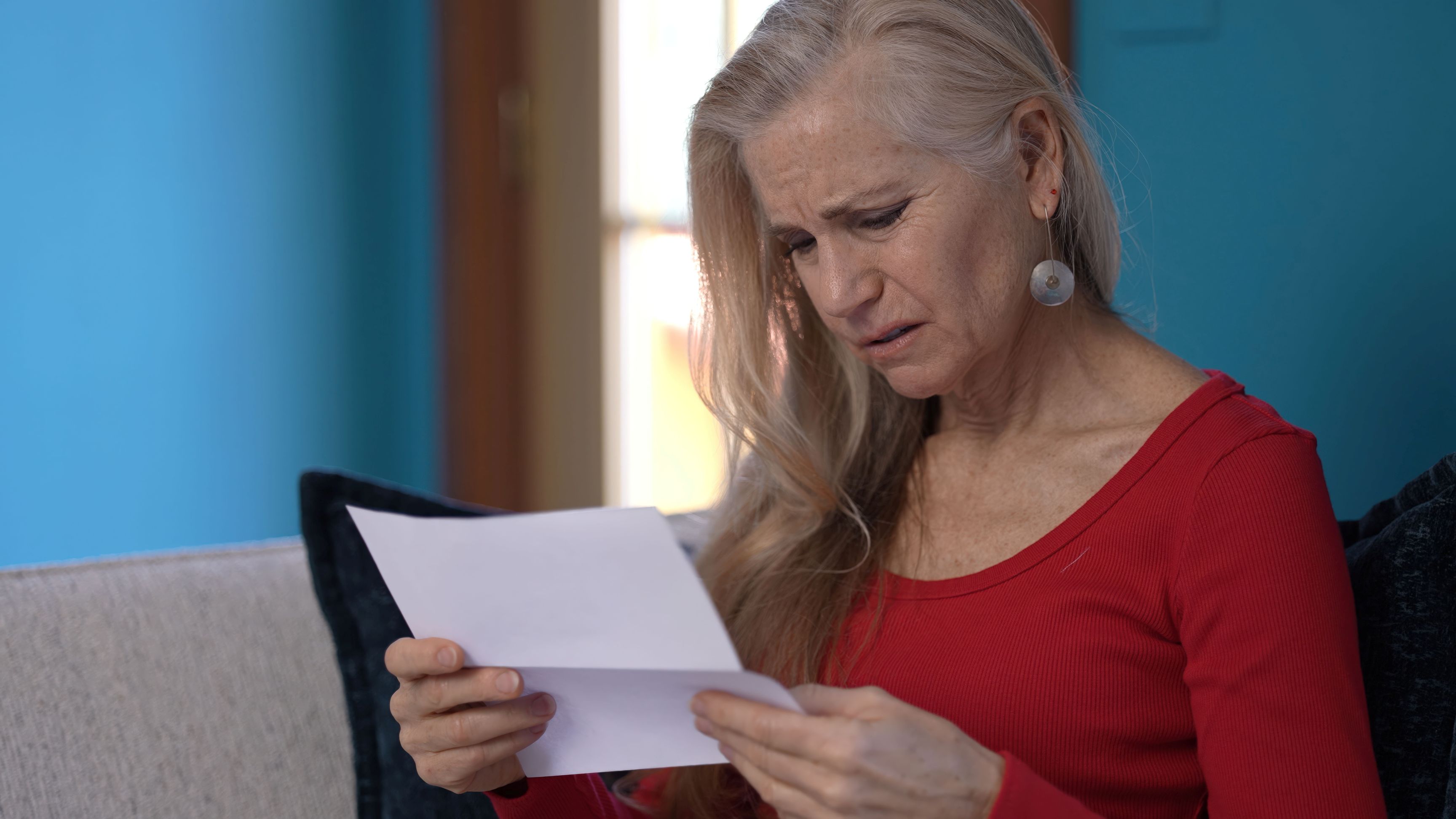 Senior woman sits on sofa reading a letter, looking concerned and frustrated.